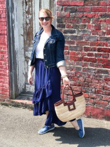 Blonde woman in casual summer skirt & sneakers look: blue ruffle skirt with jean jacket and blue slip-on sneakers posing in front of brick wall.