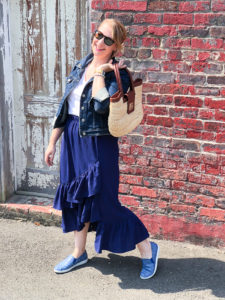 Blonde woman in casual summer skirt & sneakers look: blue ruffle skirt with jean jacket and blue slip-on sneakers posing in front of brick wall.