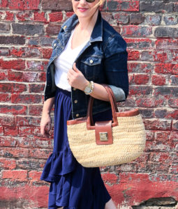 Blonde woman in casual summer skirt & sneakers look: blue ruffle skirt with jean jacket and blue slip-on sneakers posing in front of brick wall.