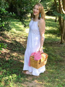 Blonde woman in white eyelet maxi dress walks in wooded area with picnic basket and champagne.
