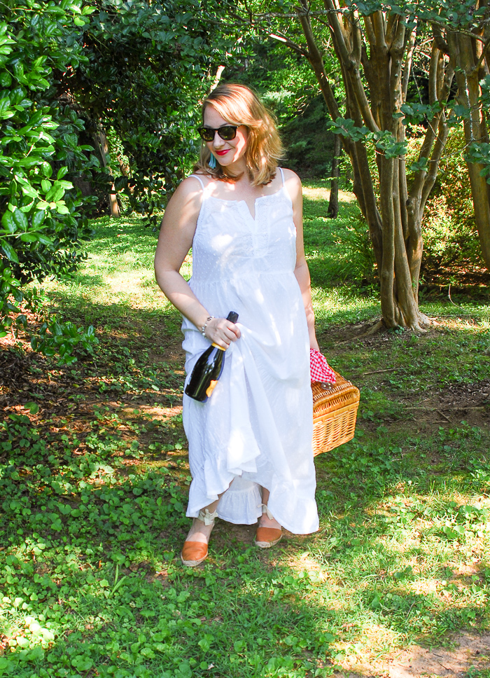 Blonde woman in white eyelet maxi dress walks in wooded area with picnic basket and champagne.