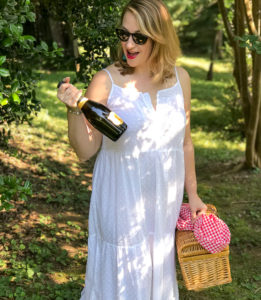 Blonde woman in white eyelet maxi dress walks in wooded area with picnic basket and champagne.