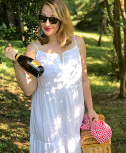 Blonde woman in white eyelet maxi dress walks in wooded area with picnic basket and champagne.