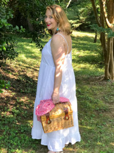 Blonde woman in white eyelet maxi dress walks in wooded area with picnic basket and champagne.