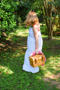 Blonde woman in white eyelet maxi dress walks in wooded area with picnic basket and champagne.