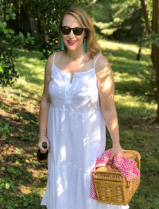 Blonde woman in white eyelet maxi dress walks in wooded area with picnic basket and champagne.