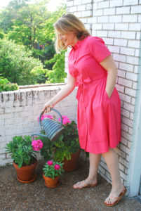 Blonde woman in pink shirtdress waters flowers on balcony