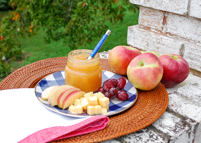 Cinnamon ginger applesauce recipe in small jar on rattan tray with sliced apples, grapes, and cheese.