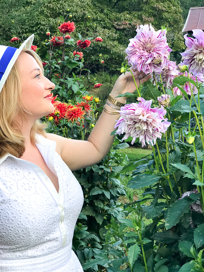 Blonde woman in white dress and hat gazes at purple dahlia