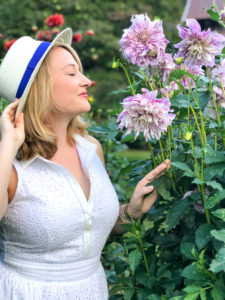 Blonde woman in white dress and hat smells purple dahlias
