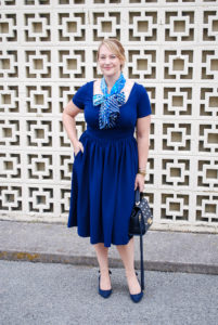 Blonde woman in navy work dress from gal meets glam with blue scarf tied in bow.