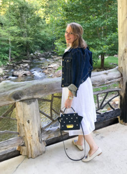Transition summer staples to fall: blonde woman on bridge overlooking stream in little white dress with denim jacket