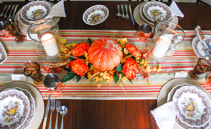 Autumn tablescape with budget friendly pumpkin centerpiece, Mason's ironstone, striped table runner, glass hurricanes, fall florals, and copper mugs.