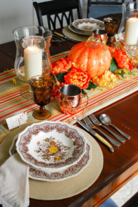 Autumn tablescape with budget friendly pumpkin centerpiece, Mason's ironstone, striped table runner, glass hurricanes, fall florals, and copper mugs.
