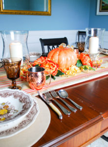 Autumn tablescape with budget friendly pumpkin centerpiece, Mason's ironstone, striped table runner, glass hurricanes, fall florals, and copper mugs.