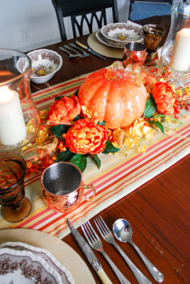 Autumn tablescape with budget friendly pumpkin centerpiece, Mason's ironstone, striped table runner, glass hurricanes, fall florals, and copper mugs.
