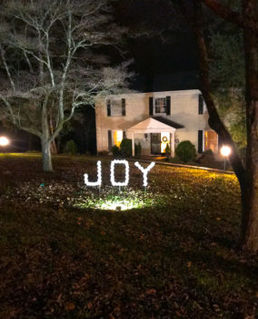 DIY outdoor Christmas decoration - PVC joy sign lit at night by spotlight in front of white brick house