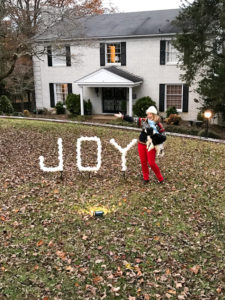 DIY outdoor Christmas decoration - PVC joy sign lit at night by spotlight in front of white brick house with woman and dog