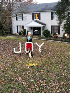 DIY outdoor Christmas decoration - PVC joy sign lit at night by spotlight in front of white brick house with woman and dog