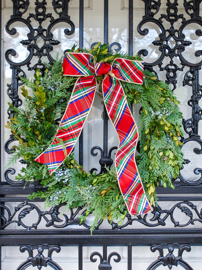 DIY Christmas wreath with fresh mixed greenery, including boxwood, cedar, and juniper hang on front door with plaid bow.
