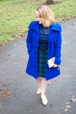 Blonde woman in dressy holiday look: black watch skirt, Draper James top, blue coat, and gold flats.