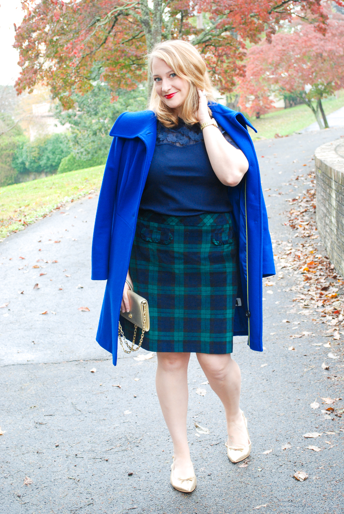 Blonde woman in dressy holiday look: black watch skirt, Draper James top, blue coat, and gold flats.
