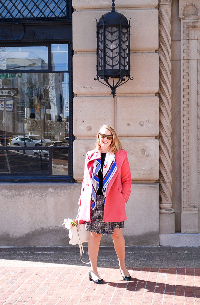 Blond woman in pink coat and tweed skirt with colorful scarf on street.