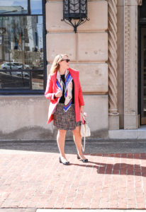 Blond woman in pink coat and tweed skirt with colorful scarf on street.