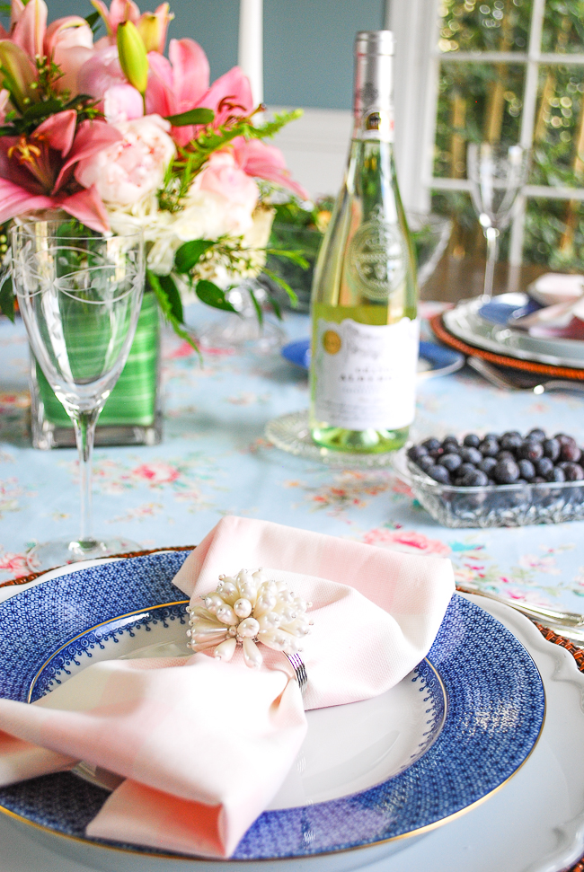 A feminine spring table setting with blue lace Mottahedeh china, rattan charger, and pink gingham napkin with pearl ring