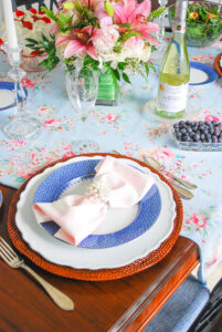 A feminine spring table setting with blue lace Mottahedeh china, rattan charger, and pink gingham napkin with pearl ring