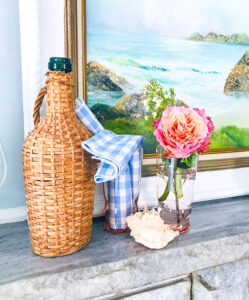 A nautical summer mantel with vintage coastal painting, wicker cask bottle, and seashells.