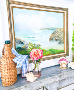 A nautical summer mantel with vintage coastal painting, wicker cask bottle, and seashells.