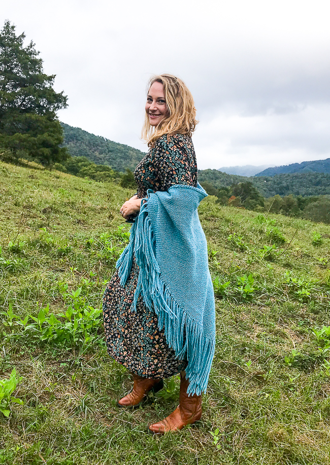 Katherine draped in blue wool shawl with floral dress and boots standing in mountain pasture walking for stress relief
