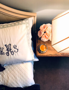 Bed with monogramed pillow and nightstand with lamp, peonies, and cup of tea