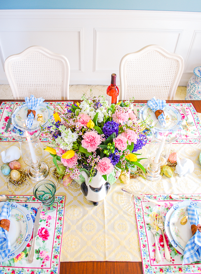 Easter tablescape set with yellow tablecloth, Pinheiro Bordallo bunny platters, Vista Alegre china, April Cornell placemats, and gingham napkins. Centerpiece is Fitz & Floyd cabbage bunny with spring florals
