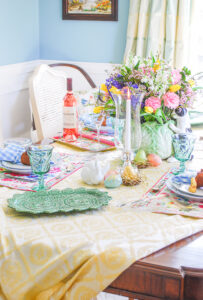 Easter tablescape set with yellow tablecloth, Pinheiro Bordallo bunny platters, Vista Alegre china, April Cornell placemats, and gingham napkins. Centerpiece is Fitz & Floyd cabbage bunny with spring florals