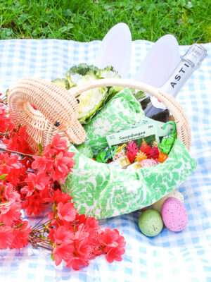Wicker swan basket for adult Easter basket inspiration filled with bottle of champagne, flower seed packets, pretty dish towels, and candy.