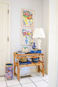 Rattan console table with marble lamp, books, catchalls, and colorful floral paintings above