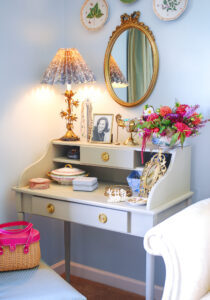 Bedroom corner with vintage vanity styled with gilt mirror, glam lamp, vintage boxes, and flowers.