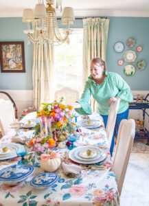 Katherine in green gingham blouse leans over to fix flowers on fall table with pastel hues, chintz tablecloth, candles, white pumpkins, and antique china.