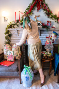 Katherine in camel sweater and gold pleated skirt lights candles on tartan meets chinoiserie traditional Christmas mantel