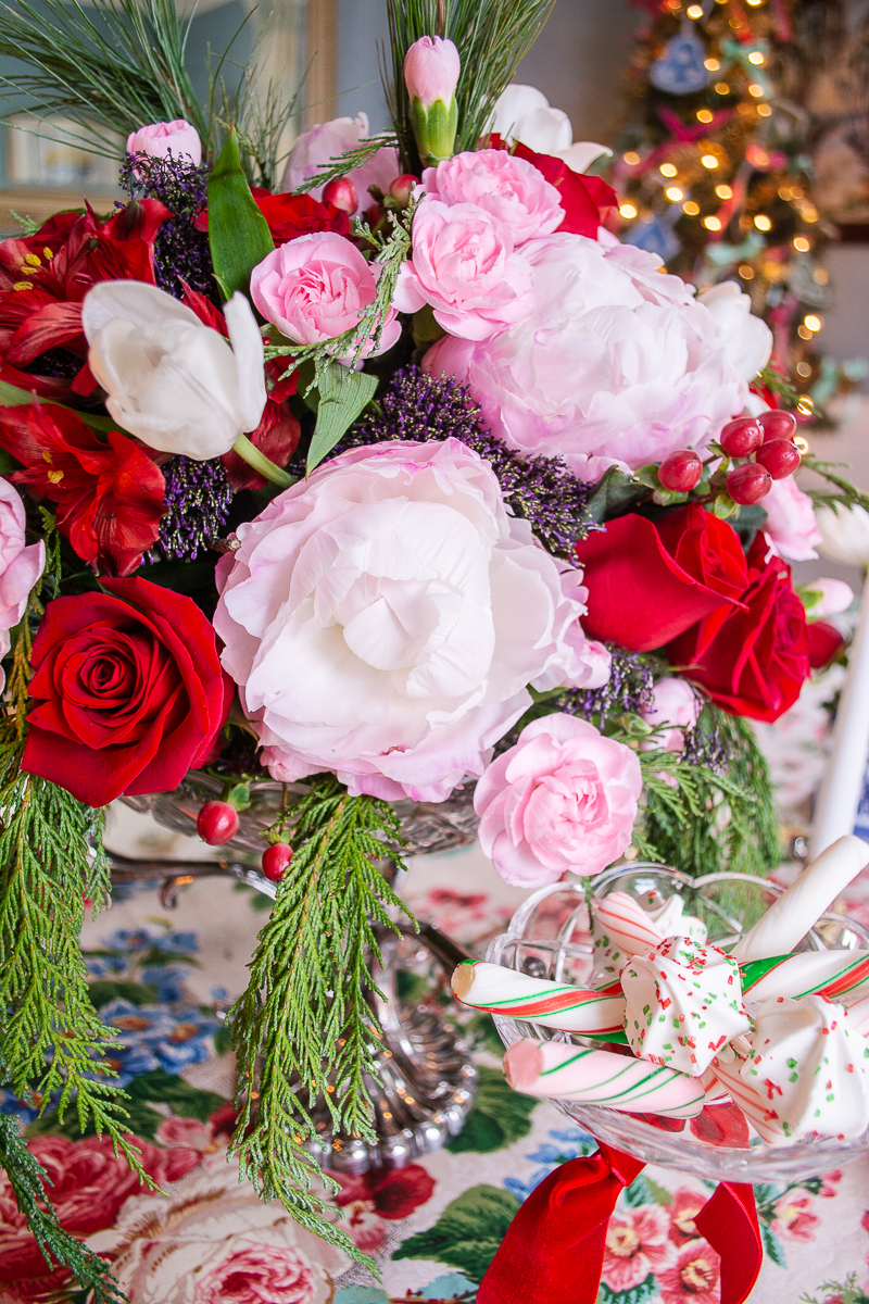 Pink peonies, red roses, pink carnations, tulips, and cedar mingle in this Christmas floral centerpiece - arranged in a silver and crystal epergne