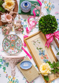 Flatlay with grandmillennial decor showcasing how to decorate with pink - boxwood topiary, pink gingham bow, botanical florals, rose medallion, wicker, Wedgwood, and needlepoint