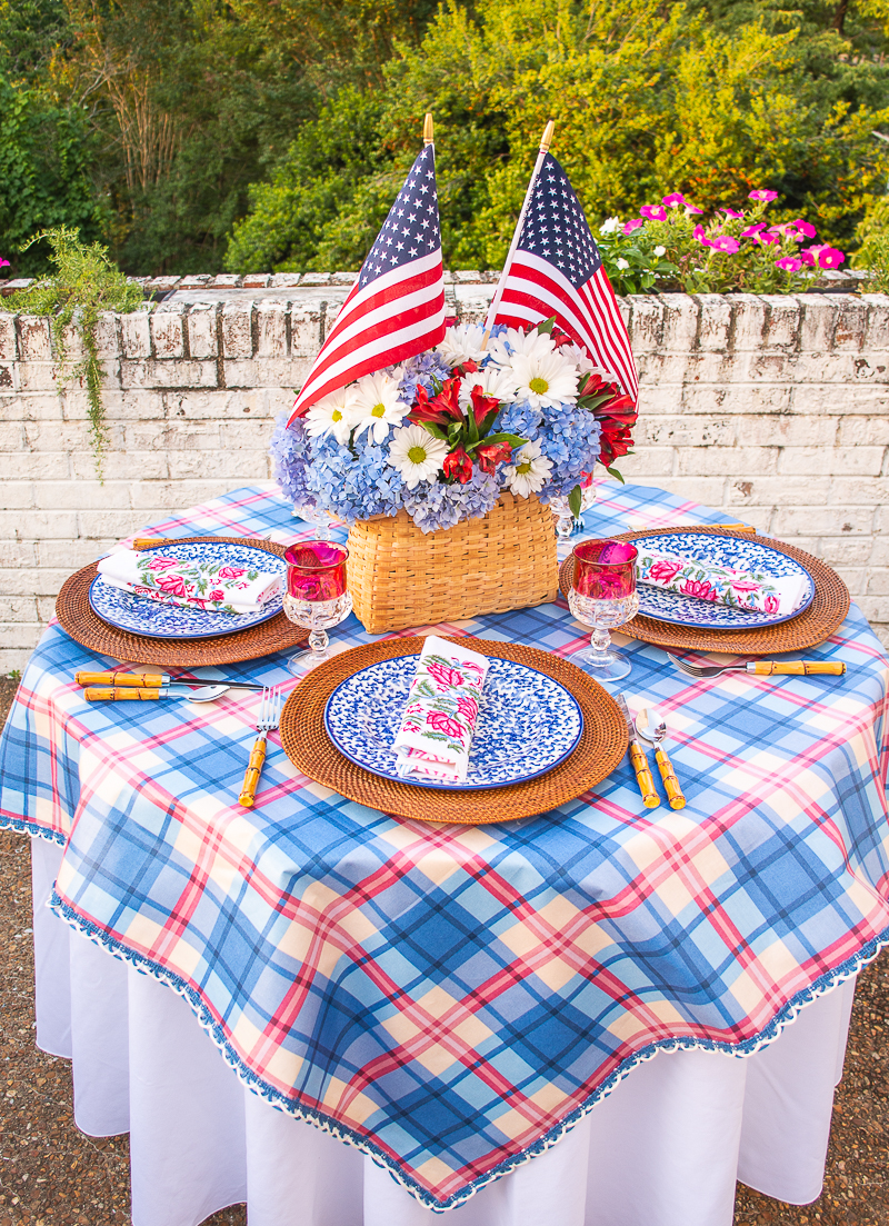 Independence day tablescape with plaid tablecloth, basket centerpiece with hydrangea and flags, splatterware dishes, and block print napkins