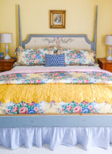 Vintage chintz bedding in yellow, pink, and blue with a gold satin quilt, and ticking stripe blue bed skirt on a painted four poster bed