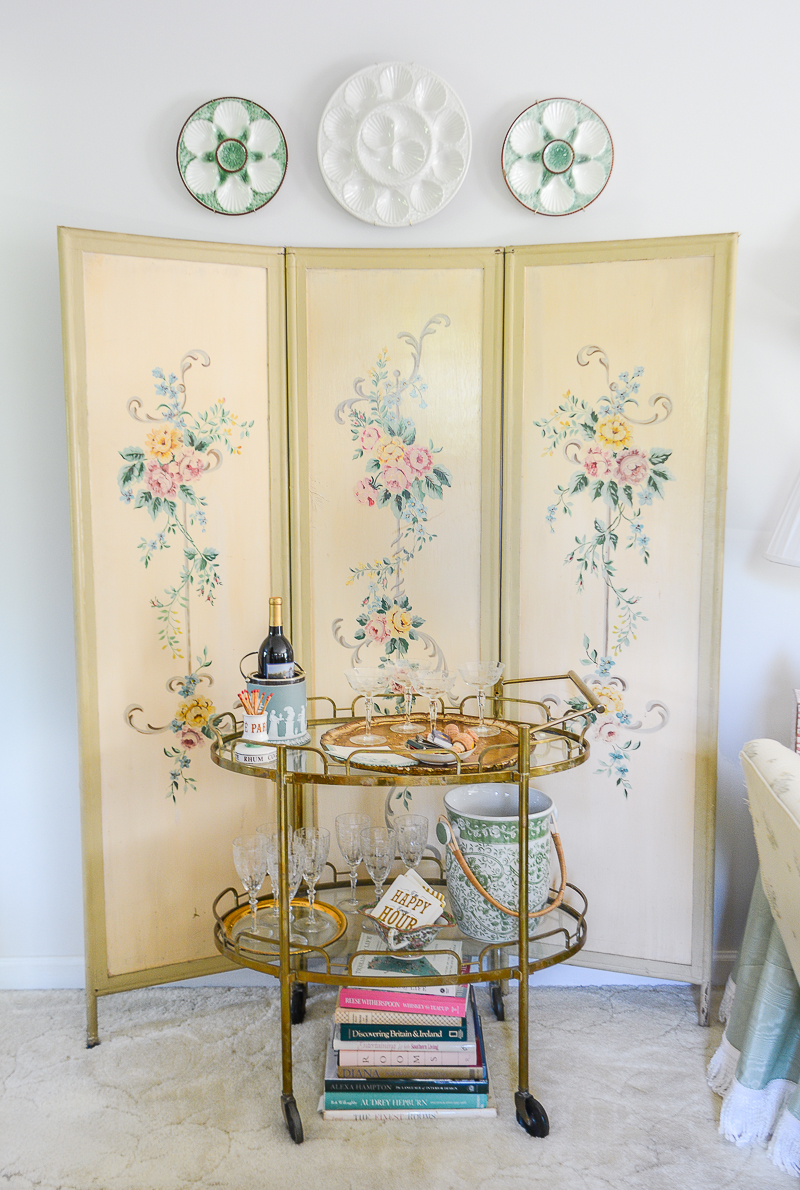 A floral folding screen accents the vintage brass bar cart with antique oyster plates above for a garden feel indoors