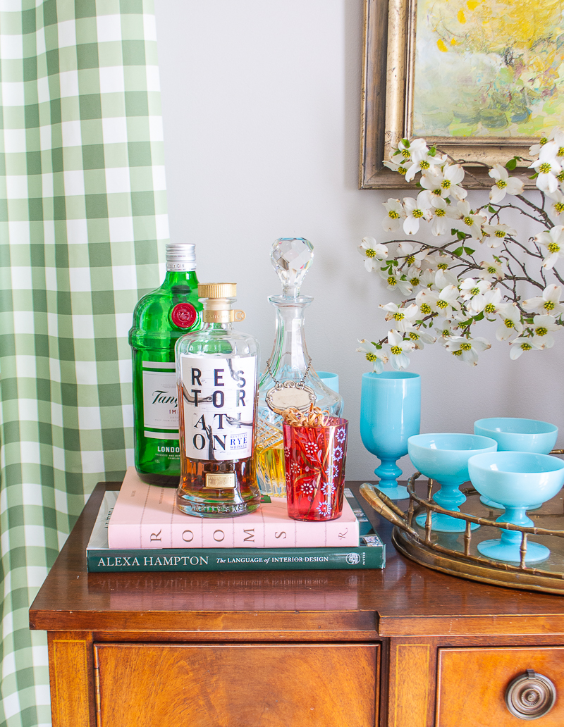 Mahogany sideboard styled as bar with aqua opaline glasses, dogwood branch, and brass tray