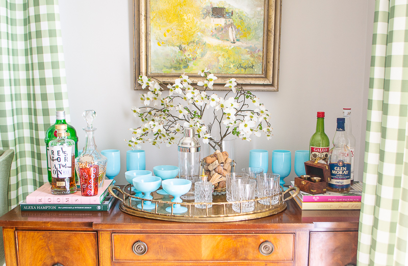 Mahogany sideboard styled as bar with aqua opaline glasses, dogwood branch, and brass tray