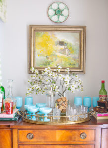 Mahogany sideboard styled as bar with aqua opaline glasses, dogwood branch, and brass tray