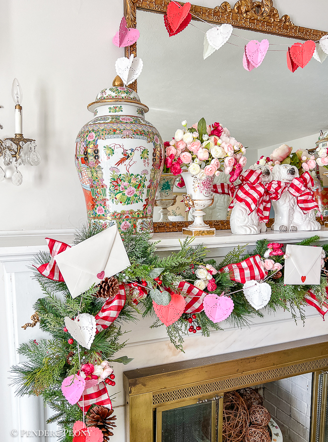A red and pink Valentine's Day mantel with crafted paper heart doily garland, gingham ribbon, love letters, and blooms
